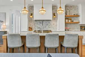 Kitchen featuring a kitchen island with sink, decorative light fixtures, backsplash, white cabinets, and light countertops