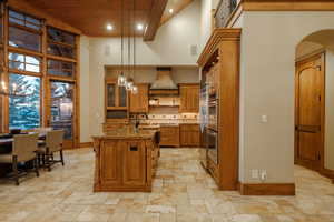 Kitchen with brown cabinets, dark stone counters, decorative light fixtures, a towering ceiling, and glass insert cabinets