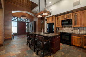 Kitchen featuring a center island with sink, black appliances, brown cabinets, a kitchen bar, and dark stone counters