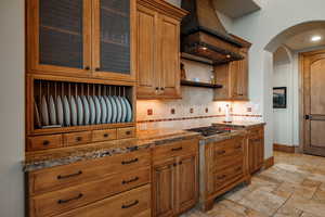 Kitchen with stainless steel gas cooktop, custom range hood, backsplash, arched walkways, and dark stone countertops
