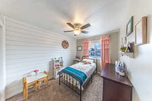 Carpeted bedroom featuring ceiling fan, wood walls, and a textured ceiling