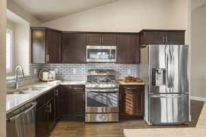 Kitchen featuring a sink, light stone counters, stainless steel appliances, dark brown cabinets, and vaulted ceiling