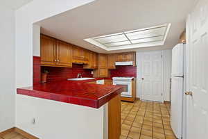 Kitchen with under cabinet range hood, white appliances, a peninsula, and tile counters