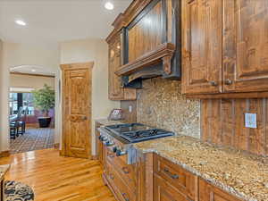 Kitchen with arched walkways, custom range hood, stainless steel gas stovetop, light wood-style floors, and backsplash