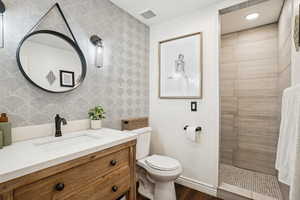 Full bathroom with visible vents, wallpapered walls, toilet, tiled shower, and vanity