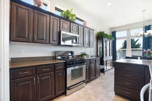 Kitchen with an inviting chandelier, appliances with stainless steel finishes, pendant lighting, dark countertops, and dark brown cabinets