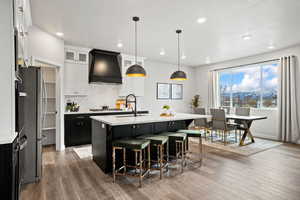 Kitchen with wood finished floors, custom exhaust hood, a kitchen island with sink, a sink, and light countertops
