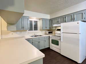 Kitchen with under cabinet range hood, a sink, a textured ceiling, dark wood-style floors, and white appliances
