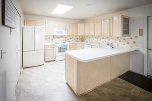 Kitchen with light brown cabinetry, a peninsula, a skylight, white appliances, and a sink