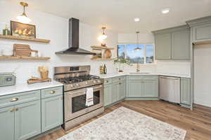 Kitchen featuring green cabinetry, open shelves, a sink, appliances with stainless steel finishes, and wall chimney range hood