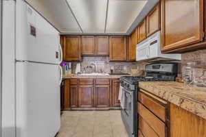 Kitchen featuring white appliances, light tile patterned floors, brown cabinetry, a sink, and tasteful backsplash