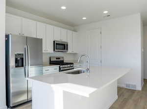 Kitchen featuring stainless steel appliances, a sink, white cabinets, an island with sink, and light wood-type flooring