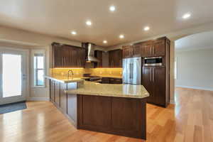 Kitchen with high quality appliances, arched walkways, a peninsula, wall chimney exhaust hood, and light wood finished floors