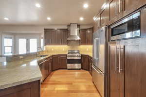 Kitchen featuring wall chimney range hood, light wood-style flooring, a peninsula, high end appliances, and a sink