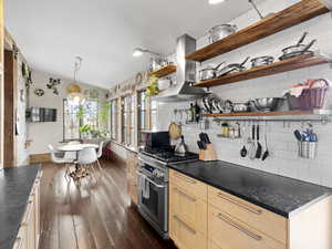 Kitchen featuring dark countertops, light brown cabinetry, high end stainless steel range oven, wall chimney exhaust hood, and open shelves