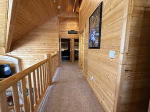 Hallway with wood walls, wood ceiling, vaulted ceiling, light colored carpet, and an upstairs landing
