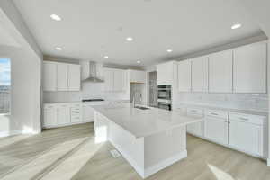 Kitchen with a center island with sink, light wood finished floors, a sink, double oven, and wall chimney exhaust hood