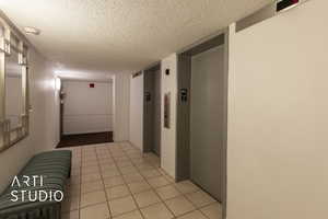 Hallway featuring elevator, light tile patterned flooring, baseboards, and a textured ceiling