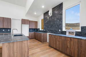 Kitchen with modern cabinets, dark stone countertops, light wood-style flooring, backsplash, and a kitchen island with sink