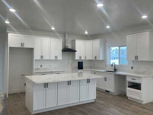 Kitchen with a kitchen island, wall chimney range hood, wood finished floors, white cabinets, and a sink
