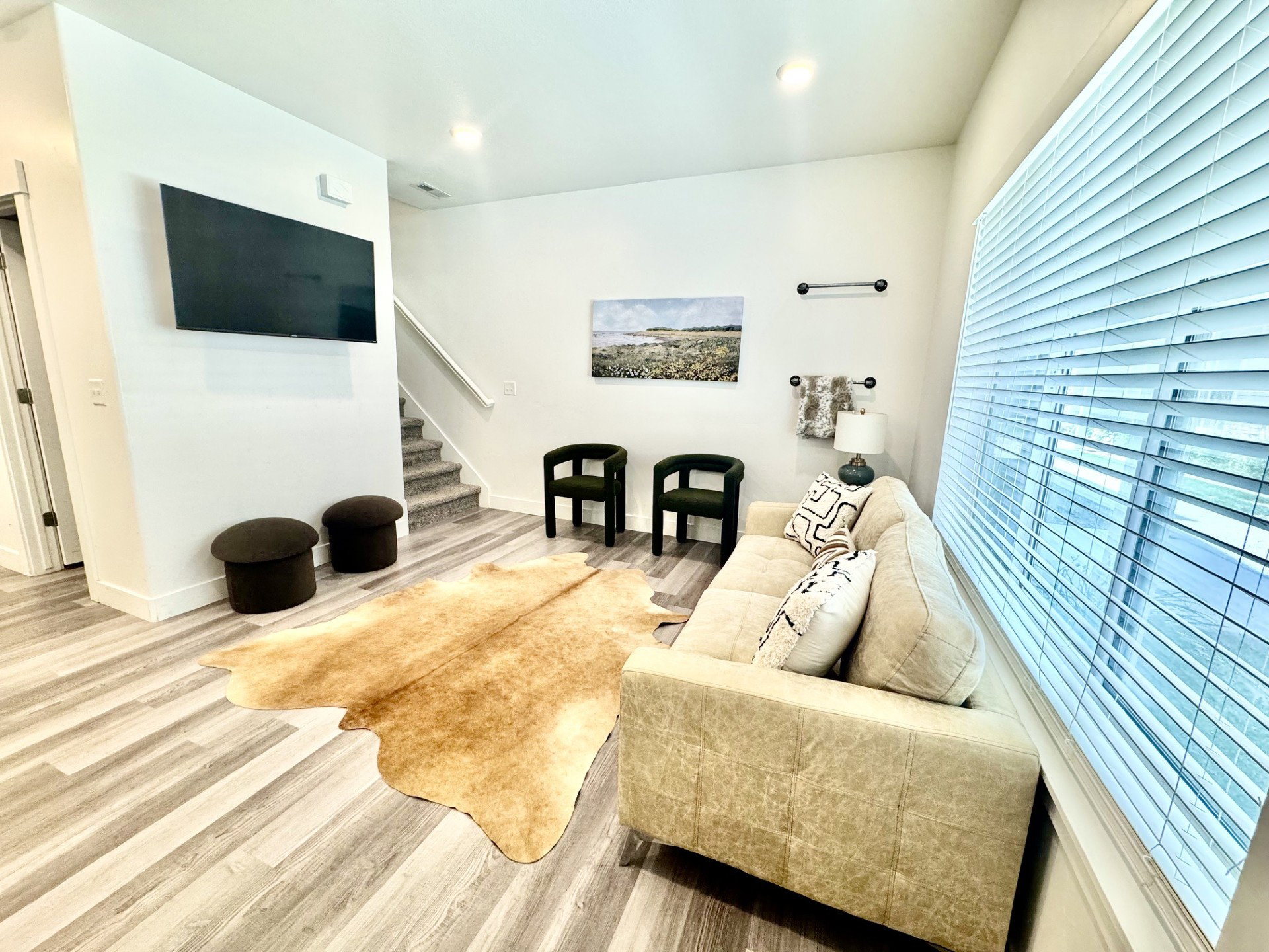 Spacious living area featuring light wood-finish flooring, a wall-mounted television, and ample natural light from a large window with blinds
