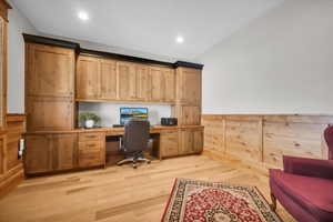 Home office with built in study area, light wood finished floors, wainscoting, vaulted ceiling, and recessed lighting
