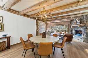 Dining room with rustic walls, a chandelier, wood finished floors, beamed ceiling, and a fireplace