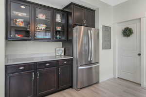 Kitchen featuring freestanding refrigerator, light wood-style floors, light countertops, glass insert cabinets, and dark brown cabinets