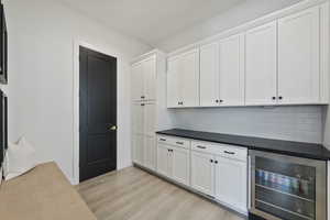 Kitchen featuring light wood-type flooring, backsplash, dark countertops, white cabinetry, and beverage refrigerator