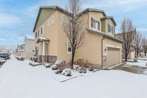 3/18 /25 snow  covered property featuring stucco siding, stone siding, a garage, and driveway