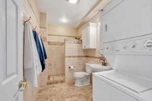 Full bath with a sink, tiled shower, tile walls, and stacked washer and clothes dryer