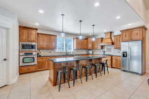 Kitchen featuring custom exhaust hood, brown cabinets, appliances with stainless steel finishes, and a kitchen island