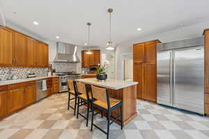 Kitchen with light floors, high end appliances, wall chimney exhaust hood, and brown cabinetry