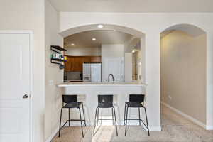 Kitchen with light colored carpet, a kitchen bar, brown cabinets, white fridge with ice dispenser, and a sink