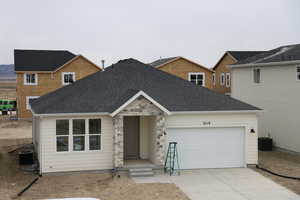 Ranch-style house featuring driveway, roof with shingles, an attached garage, stone siding, and central air condition unit