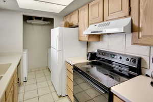Kitchen featuring light brown cabinetry, light countertops, black electric range, under cabinet range hood, and backsplash