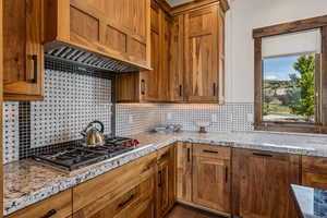 Kitchen featuring custom range hood, stainless steel gas stovetop, tasteful backsplash, and light stone countertops