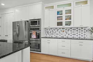 Kitchen with dark stone counters, decorative backsplash, white cabinets, light wood-style floors, and appliances with stainless steel finishes