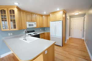 Kitchen featuring white appliances, light wood finished floors, a peninsula, light countertops, and glass insert cabinets