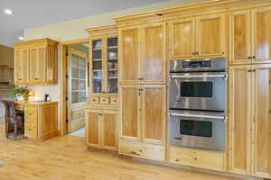 Kitchen with stainless steel double oven, glass insert cabinets, light wood-style flooring, and recessed lighting