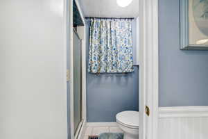 Bathroom featuring a wainscoted wall, tile patterned floors, toilet, an enclosed shower, and a textured ceiling