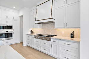 Kitchen with white cabinets, custom exhaust hood, appliances with stainless steel finishes, hardwood floors, and marble counters