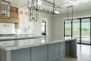 Kitchen with an island with sink, light stone counters, backsplash, light wood finished floors, and glass insert cabinets