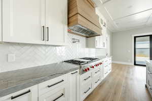 Kitchen featuring stainless steel gas cooktop, light stone counters, premium range hood, light wood-style flooring, and decorative backsplash