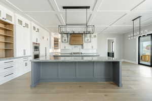 Kitchen featuring white cabinetry, a large island with sink, stainless steel double oven, open shelves, and light wood-style floors