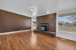 Unfurnished living room featuring baseboards, wooden walls, visible vents, a brick fireplace, and wood finished floors