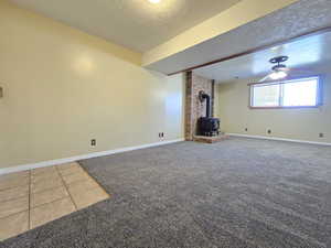 Unfurnished living room featuring a wood stove, a textured ceiling, carpet flooring, a ceiling fan, and baseboards