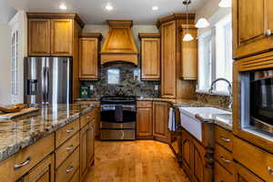 Kitchen with brown cabinets, backsplash, custom range hood, and appliances with stainless steel finishes