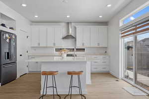 Kitchen with light countertops, refrigerator with ice dispenser, white cabinets, wall chimney exhaust hood, and a center island with sink