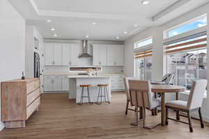 Kitchen with light countertops, a kitchen bar, light wood finished floors, white cabinets, and wall chimney exhaust hood